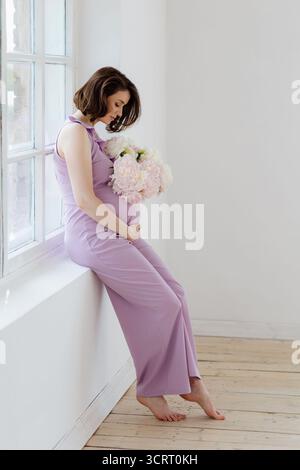 Una bella donna incinta sta accanto a una finestra, tenendo in mano un mazzo di fiori morbidi mentre indossa un vestito di lavanda. Foto Stock