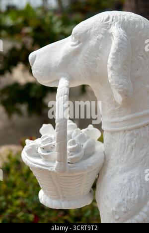 Un'affascinante scultura di cane bianco sorge in un giardino, tenendo in mano un cesto delicato pieno di fiori. La luce solare intensa mette in risalto i dettagli, creando Foto Stock
