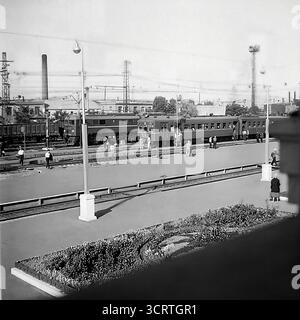 Una fotografia d'archivio in bianco e nero ad alto angolo degli anni '1970, che mostra una stazione ferroviaria affollata a Sloviansk, RSS Ucraina. La scena cattura l'essenza di un hub di trasporto industriale sovietico, con un classico treno elettrico passeggeri serie ER (elektrichka) su un binario e una locomotiva diesel con un treno merci su un altro. I passeggeri sono visti camminare sulle piattaforme sullo sfondo di un vasto paesaggio industriale con camini di fabbrica. Si tratta di un'autentica istantanea del ruolo vitale delle ferrovie nella vita quotidiana e nell'economia della regione del Donbas Foto Stock