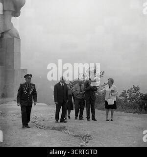 Una suggestiva fotografia d'archivio in bianco e nero degli anni '1960 mostra una delegazione ufficiale che visita il colossale Monumento ad Artem a Sviatohirsk, RSS Ucraina. Il gruppo, che include funzionari del partito e veterani di guerra decorati, cammina alla base della statua gigante. Progettato da Ivan Kavaleridze, il monumento è un capolavoro di arte sovietica famoso in tutto il mondo, eseguito in uno stile cubista/costruttivista unico. Questa immagine cattura un pellegrinaggio ufficiale verso uno dei monumenti ideologici più significativi della regione del Donbas e dell'intera URSS Foto Stock