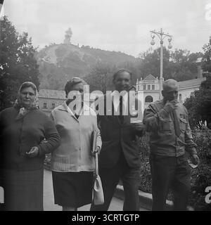 Una candida fotografia d'archivio in bianco e nero degli anni '1970, che mostra un gruppo di cittadini sovietici che si godono una passeggiata presso la stazione termale di Sviatohirsk, la RSS Ucraina. Il gruppo, che include un veterano di guerra decorato, sta passeggiando attraverso i giardini del parco. Sullo sfondo, il famoso Monumento colossale ad Artem è chiaramente visibile in cima a una collina di gesso, che si affaccia sul resort. Questa immagine nostalgica cattura un momento di tranquillo svago e turismo in una popolare città termale di epoca sovietica, con un importante punto di riferimento ideologico che forma lo sfondo della vita quotidiana Foto Stock
