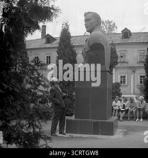 Un'autentica fotografia d'archivio in bianco e nero degli anni '1970 mostra un uomo sovietico in posa per una foto ricordo accanto a un grande monumento a Sviatohirsk, RSS Ucraina. L'uomo in tuta si erge orgogliosamente accanto al monumentale busto di una figura comunista. La scena, probabilmente ambientata sul terreno di un sanatorio o in un parco pubblico, è un tipico esempio di turismo dell'era sovietica, dove i visitatori spesso si fotografavano con punti di riferimento ideologici. Sullo sfondo, un gruppo di donne può essere visto riposare su una panchina Foto Stock