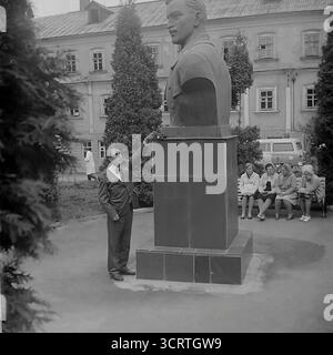 Un'autentica fotografia d'archivio in bianco e nero degli anni '1970 mostra un uomo sovietico in posa per una foto ricordo accanto a un grande monumento a Sviatohirsk, RSS Ucraina. L'uomo in tuta si erge orgogliosamente accanto al monumentale busto di una figura comunista. La scena, probabilmente ambientata sul terreno di un sanatorio o in un parco pubblico, è un tipico esempio di turismo dell'era sovietica, dove i visitatori spesso si fotografavano con punti di riferimento ideologici. Sullo sfondo, un gruppo di donne può essere visto riposare su una panchina Foto Stock