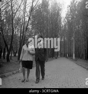 Una suggestiva fotografia d'archivio in bianco e nero degli anni '1970 mostra una coppia sovietica che fa una passeggiata romantica in un parco a Sviatohirsk, RSS Ucraina. Un uomo e una donna di mezza età camminano a braccia chiuse lungo un lungo vicolo lastricato fiancheggiato da alti pioppi. L'immagine cattura splendidamente un momento tranquillo e tranquillo di svago e di vita quotidiana in una località turistica dell'URSS durante l'era Brezhnev, evocando un potente senso di nostalgia e romanticismo senza tempo Foto Stock