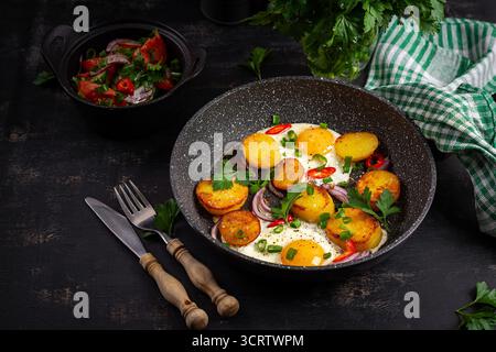 Uova e patate fritte con cipolle e peperoncino e insalata fresca su sfondo scuro. Foto Stock
