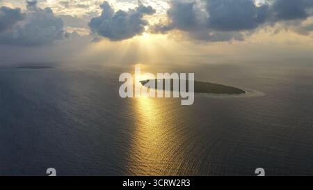 Vista aerea dei raggi dorati del sole che brillano attraverso l'oceano scuro, illuminando una piccola isola in lontananza, le Maldive. Foto Stock