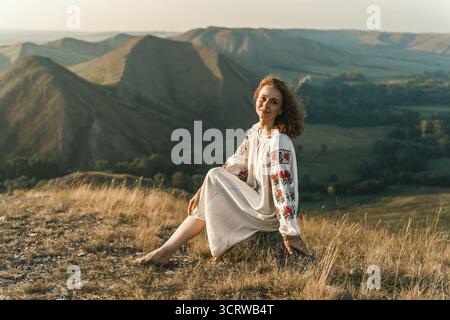 Donna in vyshyvanka siede a piedi nudi sulla roccia godendo della vista. Foto Stock
