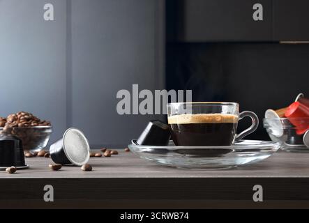 Macchina per caffè a capsule per preparare l'espresso in una tazza di vetro su un banco da cucina in legno con capsule di caffè e una cucina scura sullo sfondo. Vista frontale. Foto Stock