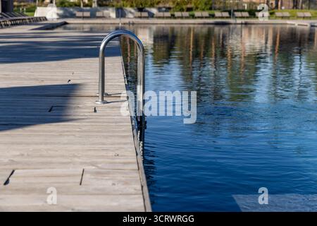Questa immagine cattura una tranquilla scena a bordo piscina caratterizzata da una scala in metallo lucido che conduce all'acqua cristallina, con pavimento in legno e riflessi morbidi Foto Stock