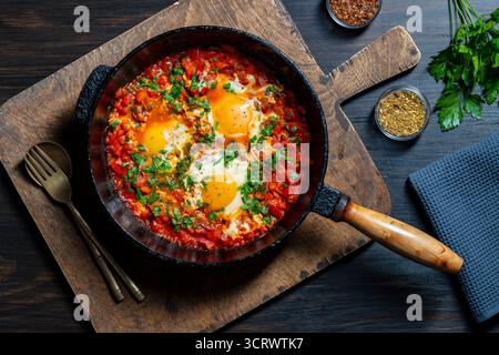 Shakshuka in padella per colazione, primo piano. Shakshuka fatto in casa con uova in camicia, cipolla, peperone, olio d'oliva, pomodoro rosso e prezzemolo in una colata Foto Stock