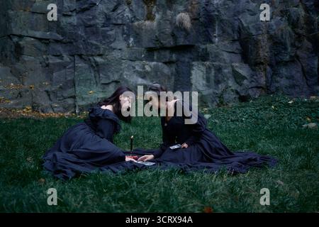 Due donne in abiti lunghi neri sedute su rocce nere ad Halloween, leggono carte di tarocchi, eseguono un rituale di strega moderna Foto Stock
