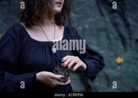 Donna con un lungo abito nero vicino a rocce nere a Halloween che regge un cristallo nero viola, che esegue un rituale di strega moderna Foto Stock