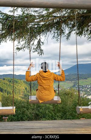 Una donna con una giacca gialla siede su un'altalena che si affaccia su una valle di montagna panoramica con foreste, villaggi e cieli nuvolosi Foto Stock