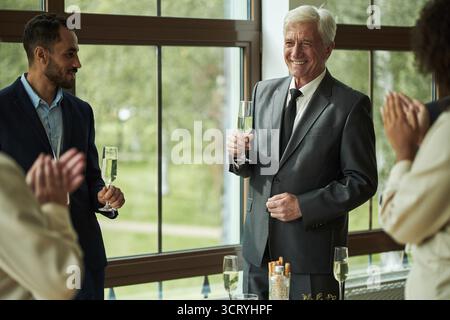 L'anziano uomo caucasico sorride e tiene in mano un calice di champagne mentre applaude i colleghi durante l'evento celebrativo, un gruppo eterogeneo di adulti impegnati in una conversazione Foto Stock