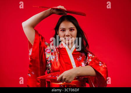 Giovane donna asiatica che indossa un tradizionale kimono rosso con motivi floreali, tiene in mano due ventole rosse e sorride alla fotocamera contro una vivace ba rossa Foto Stock