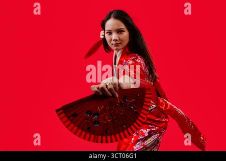 Giovane donna asiatica che indossa un tradizionale kimono rosso con motivi floreali, tiene in mano due ventole rosse e sorride alla fotocamera contro una vivace ba rossa Foto Stock