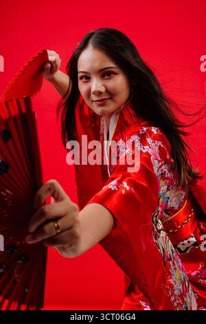 Giovane donna asiatica che indossa un tradizionale kimono rosso con motivi floreali, tiene in mano due ventole rosse e sorride alla fotocamera contro una vivace ba rossa Foto Stock