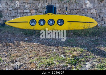 MASLINICA, CROAZIA, 08.25.2025: Una scultura subacquea di colore giallo vivo poggia su un prato e roccioso in primo piano contro un grezzo muro di pietra. Quattro oblò circolari Foto Stock