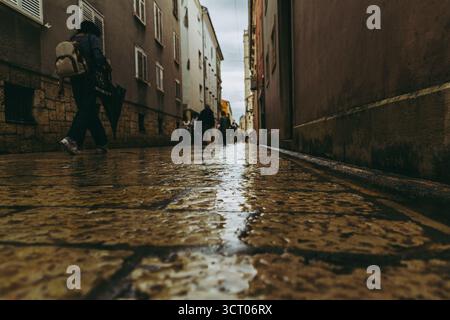 Strada bagnata in pietra a Zara, Croazia, dopo la pioggia, con pedoni sotto cieli nuvolosi e un'atmosfera tranquilla. Foto Stock