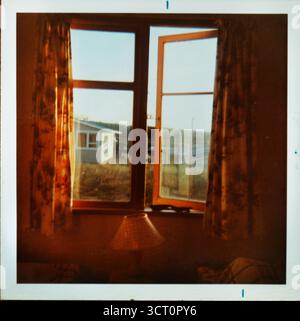 Immagine analogica di finestre con cornici in legno e vecchie tende. Si affaccia su una camera da letto in una casa sulla spiaggia di metà secolo in nuova Zelanda, con la luce del sole nel tardo pomeriggio Foto Stock