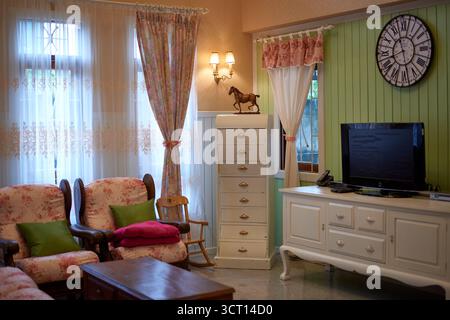 Uno spazio abitativo caldo e invitante presenta poltrone color pastello, un comò bianco e tende che filtrano la luce del sole. Un orologio nostalgico e un dondolo Foto Stock