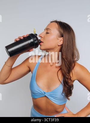 Una giovane donna con un abito atletico azzurro beve da una tazza nera. Ha i capelli lunghi e gode di un momento di idratazione in una spa fresca e pulita Foto Stock