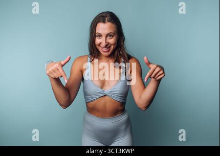 Una donna sorride brillantemente mentre indossa abbigliamento sportivo azzurro. Sta posando con le mani rivolte verso il basso, creando un'atmosfera invitante. Foto Stock