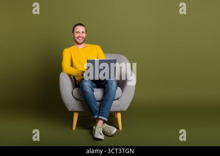 Sorridente giovane uomo in stile casual che utilizza un notebook mentre si siede su una sedia moderna su uno sfondo verde Foto Stock