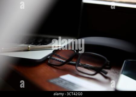Una vista ravvicinata degli occhiali da vista che si trovano accanto a una penna e a un computer portatile su una scrivania in legno lucido con riflessi morbidi. L'immagine mostra un lavoro minimalista ordinato Foto Stock