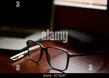 Un primo piano di occhiali da lettura appoggiati su una scrivania in legno lucido accanto a una penna dorata e a un computer portatile. La composizione crea un ufficio professionale elegante Foto Stock