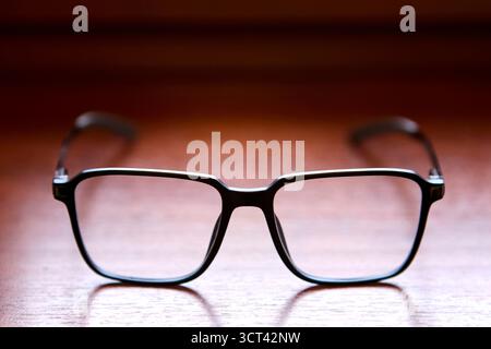 Occhiali da vista neri fotografati dalla parte anteriore, posizionati su una superficie in legno con le braccia leggermente sfocate sullo sfondo, per creare un look minimale e simulato Foto Stock