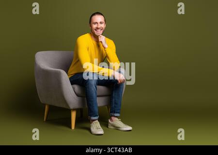 Un giovane sorridente seduto su una sedia grigia su sfondo verde, che mostra stile informale e stile amichevole Foto Stock