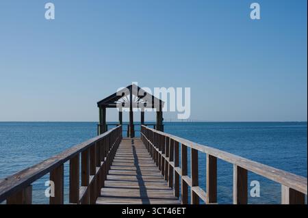Un lungo molo di legno verso un gazebo riparato si estende sulla tranquilla baia di Tampa sotto il cielo blu brillante a San Pietroburgo, Florida. Acqua tranquilla e lin pulito Foto Stock