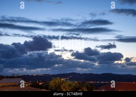 Strati di nuvole spettacolari si spostano su un paesaggio collinare ondulato al crepuscolo. Il cielo blu dell'ora fornisce uno splendido sfondo per le luci scintillanti di un ru Foto Stock