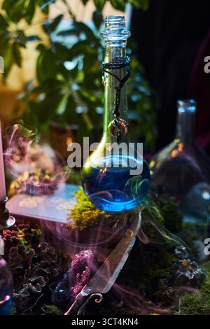 una pozione blu vibrante si trova in una bottiglia di vetro, circondata da piante verdi e luce di candela soffusa. Il fumo scorre delicatamente intorno a una scena mistica, creando un Foto Stock