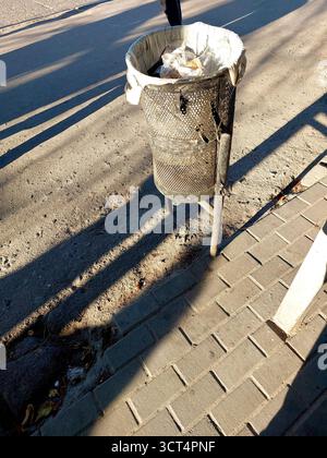 Un cestino a rete nera si trova su una strada con la luce del sole che proietta lunghe ombre. Contiene articoli scartati e si trova vicino a un marciapiede. Foto Stock