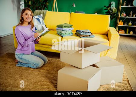 Donna sorridente che organizza gli effetti personali in un salotto colorato con scatole da imballaggio e vestiti piegati per una mossa Foto Stock