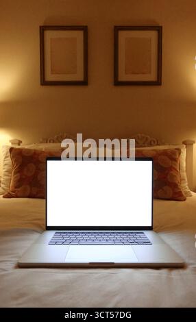 Computer portatile con schermo bianco vuoto appoggiato su un letto in un'accogliente camera da letto, con illuminazione calda e invitante. Copyspace di mockup Foto Stock
