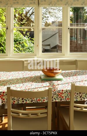 Tavolo da pranzo con tovaglia floreale e ciotola in legno, che cattura la luce naturale del sole attraverso una finestra. Campagna dei Cotswolds, Inghilterra Foto Stock