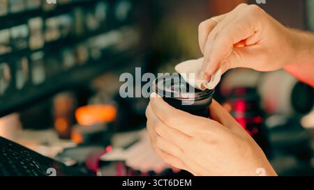 Pulizia delle mani del fotografo per la manutenzione dell'obiettivo della fotocamera. Primo piano di un uomo che pulisce l'obiettivo DSLR con un panno in microfibra alla scrivania in un moderno studio creativo, Profe Foto Stock