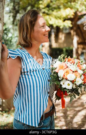 Una donna che indossa una maglia a righe sorride mentre tiene in mano un vivace bouquet di fiori in un ambiente all'aperto. La sua espressione gioiosa e la composizione floreale crea Foto Stock