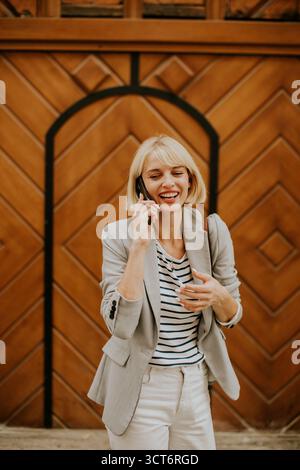 Una donna elegante si trova di fronte a una porta in legno dal design complesso e chiacchiera gioiosamente sul suo telefono. Indossa un outfit alla moda, Radiati Foto Stock