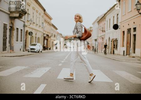 Una donna con i capelli corti passeggia lungo un incrocio in una tranquilla zona urbana. Indossa abiti casual e porta con sé una borsa, trascorrendo una piacevole giornata Foto Stock