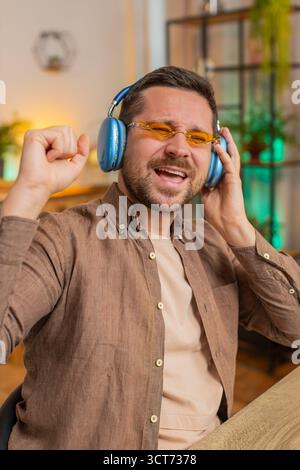 Giovane uomo d'affari felice e rilassato in ufficio che indossa cuffie e occhiali da sole ascoltando la musica da discoteca preferita e ballando. Uomo libero professionista che si rilassa, fa una pausa, festeggia. Foto Stock