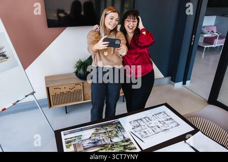 Due donne, una che indossa una camicetta rossa, sorridono con gioia mentre scattano un selfie in un ufficio moderno. I progetti e i disegni architettonici sono visibili Foto Stock