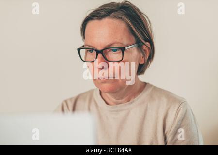 Donna adulta concentrata che indossa gli occhiali, utilizza un computer portatile nello spazio di lavoro domestico, abbigliamento casual. Messa a fuoco selettiva. Foto Stock