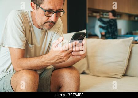 Uomo di mezza età seduto su un divano usando lo smartphone mentre la donna lavora in cucina sullo sfondo. Messa a fuoco selettiva. Foto Stock