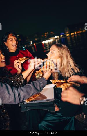 Un allegro gruppo di amici che gusta la pizza all'aperto vicino alle luci della città sul mare durante la notte. Foto Stock