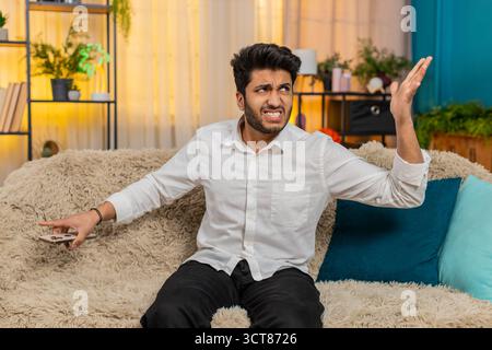 L'uomo indiano a casa sul divano afferrando lo smartphone ermeticamente, sconvolto, con rabbia buttandolo da parte dopo cattive notizie. Ragazzo arabo frustrato, infastidito, reagito emotivamente, mostrando stress, delusione, irritazione Foto Stock