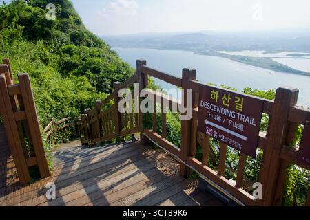 Un cartello "Descending Trail" segna la scalinata di Seongsan Ilchulbong, con ringhiere in legno che scendono lungo il ripido pendio verso la costa e la laguna Foto Stock
